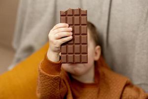 petit garçon adorable assis sur le canapé à la maison et mangeant une barre de chocolat. enfant et bonbons, confiserie de sucre. enfant savoure un délicieux dessert. enfant d'âge préscolaire avec des vêtements décontractés. photo