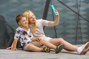 femme blonde heureuse et petit garçon assis sur la terrasse et faisant selfie sur smartphone. mère et fils passent du temps ensemble. jeune maman positive passant du temps avec son enfant mignon. s'amuser. famille. photo