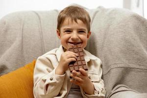 petit garçon adorable assis sur le canapé à la maison et mangeant une barre de chocolat. enfant et bonbons, confiserie de sucre. enfant savoure un délicieux dessert. enfant d'âge préscolaire avec des vêtements décontractés. émotion positive. photo