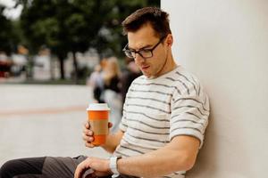 jeune homme adulte assis à l'extérieur, buvant du café et attendant quelqu'un. homme regardant sa montre et attendant sa petite amie. l'homme a une pause du travail, passe du temps à l'extérieur et essaie de se détendre. photo