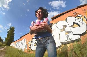 portrait d'une jeune fille émotionnelle aux cheveux noirs et aux piercings. une photo grand angle d'une fille avec des pots de peinture aérosol dans les mains sur un fond de mur de graffitis. un portrait moderne d'un objectif fisheye