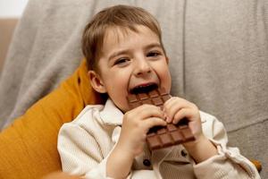 petit garçon adorable assis sur le canapé à la maison et mangeant une barre de chocolat. enfant et bonbons, confiserie de sucre. enfant savoure un délicieux dessert. enfant d'âge préscolaire avec des vêtements décontractés. émotion positive. photo