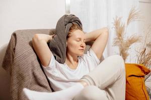 jeune femme blonde avec des vêtements décontractés, une serviette sur la tête assise sur un canapé et se reposant. détendez-vous après avoir pris une douche. prendre soin de soi, du temps pour soi. détente à la maison. concept de femmes, de beauté et d'hygiène. photo