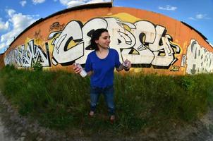 portrait d'une jeune fille émotionnelle aux cheveux noirs et aux piercings. une photo grand angle d'une fille avec des pots de peinture aérosol dans les mains sur un fond de mur de graffitis. un portrait moderne d'un objectif fisheye