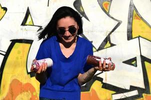 portrait d'une jeune fille émotionnelle aux cheveux noirs et aux piercings. photo d'une fille avec des pots de peinture aérosol dans les mains sur un fond de mur de graffiti. le concept de street art et l'utilisation de peintures aérosols