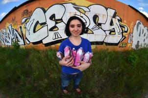 portrait d'une jeune fille émotionnelle aux cheveux noirs et aux piercings. une photo grand angle d'une fille avec des pots de peinture aérosol dans les mains sur un fond de mur de graffitis. un portrait moderne d'un objectif fisheye