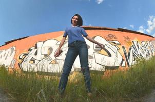 portrait d'une jeune fille émotionnelle aux cheveux noirs et aux piercings. une photo grand angle d'une fille avec des pots de peinture aérosol dans les mains sur un fond de mur de graffitis. un portrait moderne d'un objectif fisheye