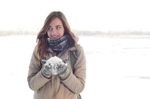 une jeune et joyeuse fille caucasienne dans un manteau marron tient une boule de neige à l'arrière-plan d'une ligne d'horizon entre le ciel et un lac gelé en hiver photo