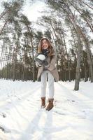 une jeune et joyeuse fille caucasienne en manteau marron tient une boule de neige dans une forêt enneigée en hiver. photo fisheye