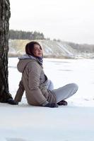 une jeune fille de race blanche dans un manteau marron est assise près d'une falaise à l'arrière-plan d'une ligne d'horizon entre le ciel et un lac gelé en hiver photo