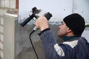 un ouvrier âgé perce un trou dans un mur en polystyrène pour l'installation ultérieure d'une cheville de renfort en plastique. créer des trous dans le mur avec une perceuse. réchauffement de la façade du bâtiment photo