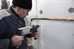 un ouvrier âgé perce un trou dans un mur en polystyrène pour l'installation ultérieure d'une cheville de renfort en plastique. créer des trous dans le mur avec une perceuse. réchauffement de la façade du bâtiment photo