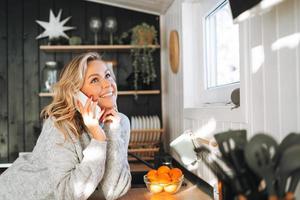 jeune femme aux cheveux bouclés blonds en pull gris utilisant un téléphone portable dans la cuisine de la maison