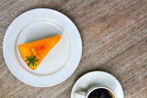 vue de dessus morceau de gâteau orange dans un plat blanc ou une assiette et une tasse blanche de café noir sur fond bois avec espace de copie. mise à plat de nourriture sucrée ou dessert sur une table ou un mur en bois. ensemble de collations pour manger. photo