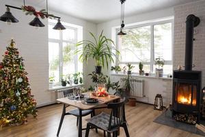 l'intérieur festif de la maison est décoré pour noël et le nouvel an dans un style loft avec poêle noir, cheminée, arbre de noël. chambre studio chaleureuse avec table dressée, feu de bois, cosy et chauffage d'accueil photo