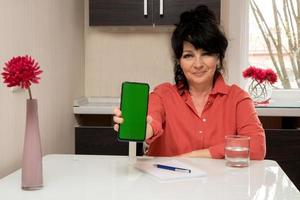 belle femme âgée est assise à la table à la maison et tient le téléphone avec un écran vert photo