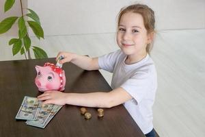 une petite fille mignonne est assise à une table sur laquelle des billets et des pièces sont disposés et met de l'argent dans une tirelire, littératie financière photo