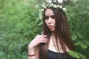 portrait d'une jeune fille émotive avec une couronne de fleurs sur la tête et des ornements brillants sur le front. jolie brune posant dans une belle forêt en plein essor pendant la journée par beau temps photo