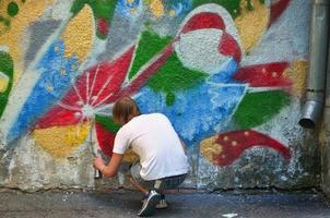 photo d'un jeune homme en short en jean et chemise blanche. le gars dessine sur le mur de graffitis un dessin avec des peintures aérosols de différentes couleurs. le concept de hooliganisme et de dommages aux biens