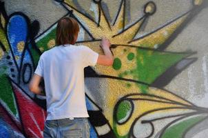 photo d'un jeune homme en short en jean et chemise blanche. le gars dessine sur le mur de graffitis un dessin avec des peintures aérosols de différentes couleurs. le concept de hooliganisme et de dommages aux biens