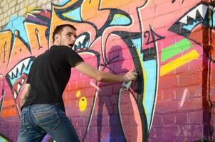 jeune graffeur avec sac à dos et masque à gaz sur son cou peint des graffitis colorés dans des tons roses sur un mur de briques. street art et processus de peinture contemporaine photo