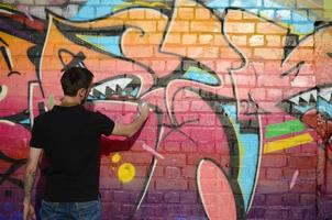 jeune graffeur avec sac à dos et masque à gaz sur son cou peint des graffitis colorés dans des tons roses sur un mur de briques. street art et processus de peinture contemporaine photo