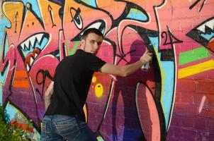 jeune graffeur avec sac à dos et masque à gaz sur son cou peint des graffitis colorés dans des tons roses sur un mur de briques. street art et processus de peinture contemporaine photo