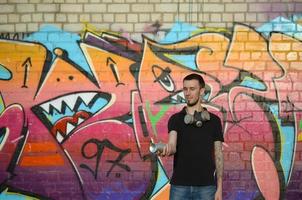 jeune graffeur caucasien en t-shirt noir avec aérosol argenté peut près de graffitis colorés dans des tons roses sur le mur de briques. street art et processus de peinture contemporaine photo
