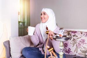 femme musulmane fumant la chicha à la maison et buvant du café ou du thé. jeune femme musulmane appréciant tout en fumant du narguilé. fille arabe fumant le narguilé photo