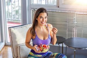 jeune et belle femme appréciant une salade saine assise à la maison avec le soleil derrière elle. concept d'aliments sains belle femme en forme mangeant une salade saine après l'entraînement de remise en forme photo