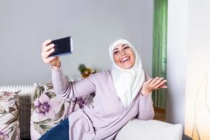 jeune femme musulmane ayant un appel vidéo via un téléphone mobile intelligent à la maison. femme musulmane souriante et heureuse assise sur un canapé, un canapé et utilisant un smartphone dans le salon à la maison, apprenant la langue, appel vidéo.