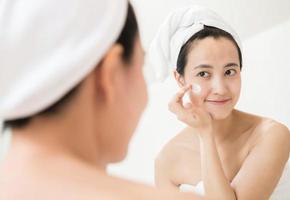 heureuse jeune femme asiatique appliquant des lotions pour le visage tout en portant une serviette et en touchant son visage dans la salle de bain photo