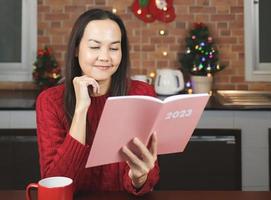 femme asiatique portant un pull en tricot rouge assis à table avec une tasse de café rouge dans la cuisine décorée d'un sapin de noël, souriant en lisant le planificateur 2023. photo