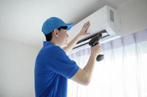 un jeune technicien asiatique homme de service portant un uniforme bleu vérifiant, nettoyant le climatiseur à la maison photo