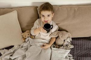 adorable petit garçon buvant du thé chaud sur le lit à la maison et se relaxant. enfant au repos, enveloppé dans une couverture, avec une tasse dans sa chambre. intérieur et vêtements aux couleurs naturelles de la terre. environnement cosy. photo