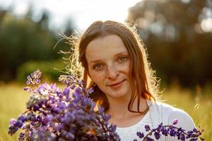 portrait en gros plan d'une belle jeune femme rousse aux taches de rousseur, vêtue d'une robe blanche, posant dans la nature. fille aux cheveux rouges tenant des fleurs. beauté naturelle. diversité, singularité individuelle. photo