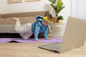 petit garçon caucasien faisant des exercices de fitness à la maison. enfant s'amusant pendant le sport. l'enfant fait de la formation, regarde l'ordinateur. sport, yoga en ligne. entraînement dans l'appartement. mode de vie sain. photo
