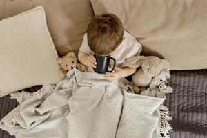 petit garçon malade buvant du thé chaud sur le lit à la maison. malade, enfant malade enveloppé dans une couverture, avec une tasse dans sa chambre. saison de la grippe. intérieur et vêtements aux couleurs naturelles de la terre. environnement cosy. photo