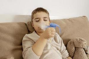 petit garçon malade avec inhalateur pour le traitement de la toux. enfant malade faisant de l'inhalation sur son lit. saison de la grippe. intervention médicale à domicile. intérieur et vêtements aux couleurs naturelles de la terre. environnement cosy. photo