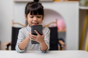 jolie petite fille utilise un smartphone tout en étant assise sur le canapé du salon. enfant surfant sur internet sur un téléphone portable, naviguant sur internet et regardant des dessins animés en ligne à la maison photo