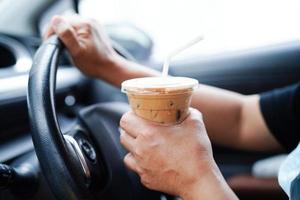 une conductrice asiatique tient une tasse de café glacé et du pain sandwich pour manger et boire en voiture, dangereuse et risque un accident. photo