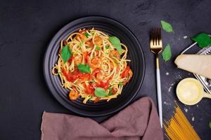 délicieuses pâtes spaghetti aux crevettes et fromage servies sur une assiette noire. avec légumes, sauce tomate italienne et épices disposés sur une table en bois, fond noir, vue de dessus photo