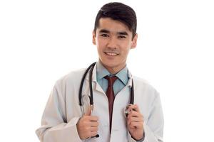 Portrait of young happy brunette man doctor in white uniform smiling on camera avec stéthoscope sur son cou isolé sur fond vide photo