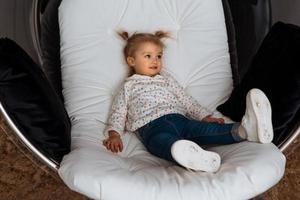 drôle de petite fille est assise sur une chaise blanche et regarde de côté photo