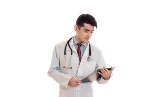 jeune homme brune sérieuse médecin en uniforme avec stéthoscope sur ses épaules prendre des notes isolé sur fond blanc photo