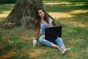 belle brune souriante est assise sur l'herbe verte avec un ordinateur portable photo