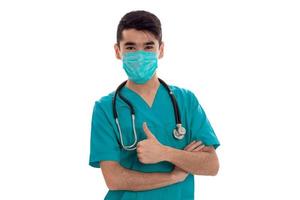 Médecin de sexe masculin en uniforme et masque avec stéthoscope montrant les pouces vers le haut isolé sur fond blanc photo