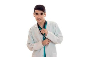 Cheerful young brunette male doctor with stethoscope en uniforme regardant la caméra isolé sur fond blanc photo