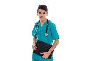 Closeup portrait de jeune beau médecin homme brune en uniforme bleu avec stéthoscope et planche dans les mains en regardant la caméra et souriant isolé sur fond blanc photo