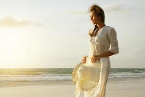 femme vêtue d'une belle robe blanche marche sur la plage au coucher du soleil photo